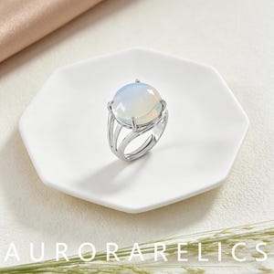 May include: A silver-toned ring featuring a large, round, smooth, translucent cabochon stone with a milky white and iridescent sheen. The band is constructed with multiple thin, parallel metal segments that meet at the base of the setting.