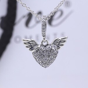 May include: A silver-toned pendant necklace featuring a heart-shaped charm adorned with small, clear gemstones. The heart has small wings on either side. The necklace chain is delicate and silver. The background is blurred.