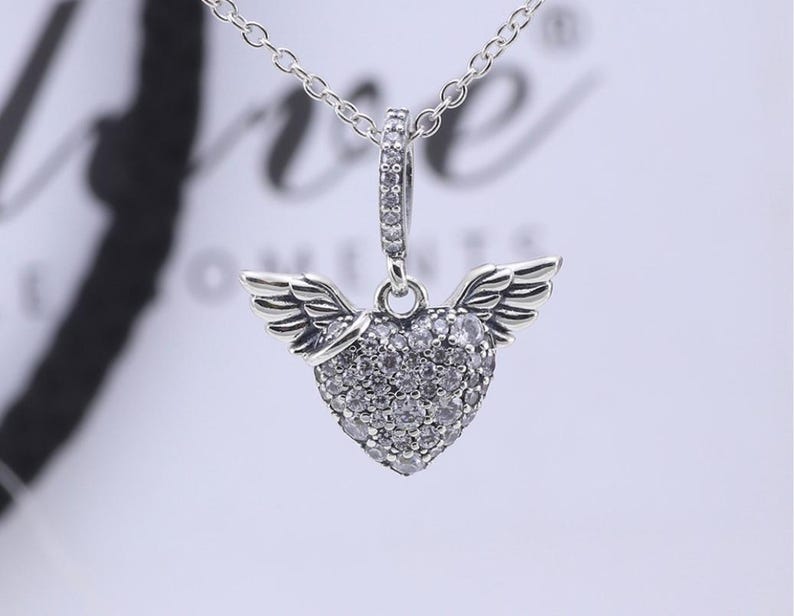 May include: A silver-toned pendant necklace featuring a heart-shaped charm adorned with small, clear gemstones. The heart has small wings on either side. The necklace chain is delicate and silver. The background is blurred.