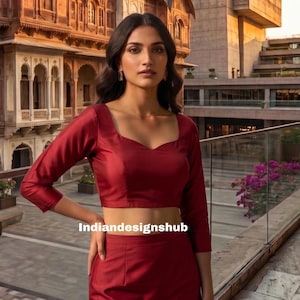 May include: A burgundy two-piece outfit featuring a square-neck, long-sleeve top and matching high-waisted skirt. The outfit is made of a smooth, shiny fabric. The text "indiandesignshub" is visible on the bottom of the image.