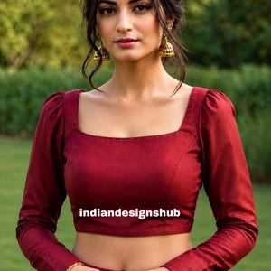 May include: A deep red long-sleeved blouse with a square neckline and puffed shoulders. The garment features the text "indiandesignshub" printed across the center. The blouse is paired with gold-toned jhumka earrings, a gold bracelet, and rings.
