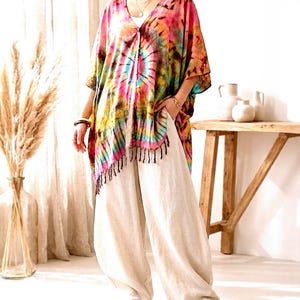May include: A tie-dye poncho featuring a vibrant mix of pink, yellow, green, and blue colors with a V-neckline and black fringe trim along the hem, paired with loose-fitting, wide-leg beige trousers and simple open-toe sandals.