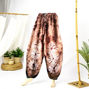 May include: Brown tie-dye pants with white circular designs. The pants have a drawstring waist and a relaxed fit. The fabric has a marbled effect with shades of brown and white. The pants are displayed on a white background.