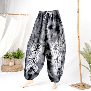 May include: Tie-dye harem pants in shades of gray and white. The pants feature a drawstring waist and a loose, comfortable fit. White circular patterns are printed on the front of the pants. The pants are made of a lightweight fabric, perfect for casual wear.