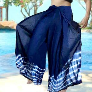 May include: Flowing navy blue palazzo pants with a tie-dye pattern in white and blue at the hem. The wide-leg pants are made of a lightweight fabric, perfect for warm weather. The pants are shown against a blurred background of water and trees.