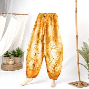 May include: Mustard yellow tie-dye pants with a drawstring waist. The pants feature a unique pattern of white circular designs. The loose-fitting pants are made of a lightweight fabric, perfect for casual wear.