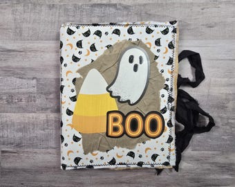 May include: A white fabric book with a black and white Halloween print. The book has a ghost, a candy corn, and the word "BOO" on the front. The book has black fabric ties.