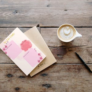 May include: A notepad with the words "To Do Liste" and pink boxes for notes, a cup of coffee with latte art, a pen, and a brown envelope on a wooden surface. The notepad has pink floral accents.