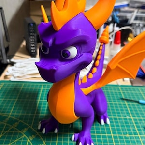 May include: A 3D-printed figurine of a purple dragon with orange detailing. The dragon features large horns, wings, and a detailed design. The figurine is on a green cutting mat.