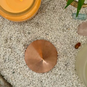 Könnte beinhalten: Ein kupferfarbener, kreisförmiger Kerzendeckel liegt auf einem Bett aus kleinen, weißen Kieselsteinen. Der Deckel hat eine konische Form mit einem kleinen Schlitz. Eine brennende Kerze in einem Glas ist im Hintergrund zu sehen, zusammen mit einer kleinen Sukkulentenpflanze.