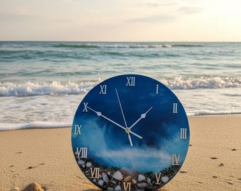 Puede incluir: Un reloj de pared redondo con un diseño azul con temática oceánica, números romanos blancos y manecillas. El reloj está colocado en una playa de arena con el océano al fondo. La esfera del reloj tiene un diseño de olas y pequeñas piedras en la parte inferior.