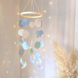 Puede incluir: Móvil hecho a mano con un anillo de madera y formas circulares iridiscentes azules y blancas colgantes. El móvil también incluye pequeñas plumas blancas y está suspendido por una cuerda.