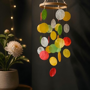 Peut inclure: Mobile décoratif avec des disques irisés colorés rouges, jaunes, verts et blancs. Le mobile est suspendu à un anneau en bois. En arrière-plan, un mur sombre et une fleur blanche en pot sont visibles.