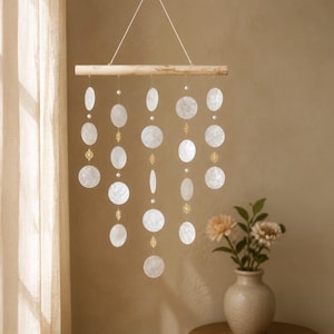 May include: A hanging mobile featuring a horizontal light-coloured wooden rod suspended by a white string. Five vertical strands hang from the rod, decorated with circular white shell discs, small white beads, and intricate gold-toned metal spacers.