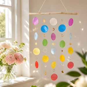 May include: A hanging mobile featuring a horizontal wooden branch suspending multiple vertical strands. These strands contain circular, translucent, multi-coloured discs in shades of blue, green, yellow, red, and purple, interspersed with small gold-toned metal charms in shapes like suns, moons, and geometric outlines.