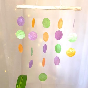 Puede incluir: Móvil hecho a mano con recortes de papel redondos y coloridos en tonos morado, verde y naranja. Los círculos están suspendidos de un pasador de madera natural con una cuerda blanca. El móvil cuelga frente a una cortina blanca.