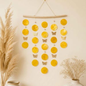 Puede incluir: Un colgante de pared con cinco hilos verticales suspendidos de una rama de madera horizontal. Los hilos consisten en discos circulares amarillos y pequeños recortes de mariposas doradas, acentuados con pequeñas cuentas blancas.