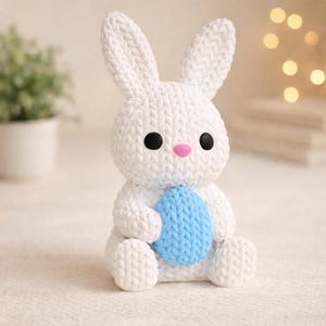 May include: A white knitted bunny figurine with black eyes and a pink nose, holding a light blue egg. The bunny has long ears and is sitting on a soft, textured surface. Decorative Easter-themed item.