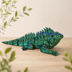 May include: A detailed, articulated iguana figurine in shades of green and blue, with a textured, metallic finish. The reptile is posed on a wooden surface, showcasing its spiky dorsal crest and segmented body. A decorative item.