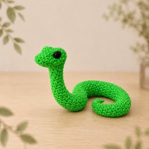 May include: A bright green crocheted snake toy with a textured yarn surface and two small, round, solid black eyes. The snake is positioned in a coiled shape with its head raised, showcasing the intricate stitch pattern of the handmade craft.