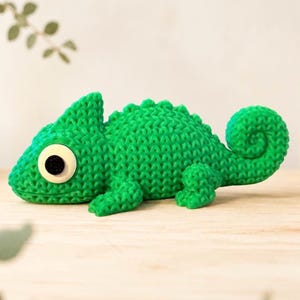 May include: A bright green chameleon figurine with a textured, knitted-style surface. The creature features a large, round, cream-coloured eye with a black pupil, a pointed crest along its back, and a curled tail. The body has a uniform, repeating pattern.