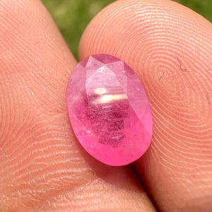 Puede incluir: Una gema rosa vibrante de forma ovalada con una superficie facetada que refleja la luz. La piedra muestra una calidad translúcida con tonos magenta profundos concentrados hacia el centro, exhibiendo un acabado pulido y un corte simétrico.