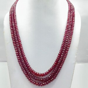 May include: A triple-strand necklace composed of small, faceted, deep red ruby beads. The beads are uniform in size and shape, creating a layered appearance with a consistent, reflective texture throughout the three strands of the necklace.