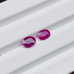 Puede incluir: Dos rubíes rojos oscuros de corte ovalado se exhiben sobre una superficie blanca. Las piedras preciosas tienen un aspecto facetado, captando la luz y mostrando su rico color. Los rubíes son de un tono rojo vibrante y saturado.