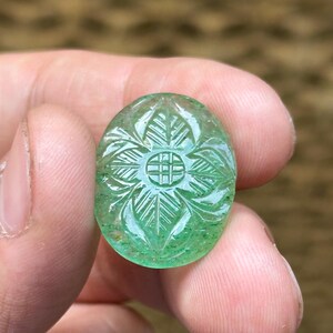 Może przedstawiać: Owalny, rzeźbiony zielony kamień szlachetny z kwiatowym wzorem. Kamień szlachetny ma centralny okrągły wzór otoczony szczegółowymi rzeźbami w kształcie liści. Powierzchnia kamienia jest wypolerowana, a kolor to żywy zielony.