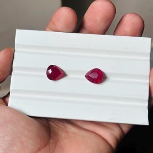 Peut inclure: Deux pierres précieuses rouges en forme de poire sont présentées sur une surface blanche. Les gemmes facettées captent la lumière, révélant leur couleur riche. Ce sont probablement des rubis, parfaits pour la joaillerie.