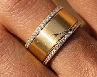Puede incluir: Un anillo de banda ancha de oro con un acabado mate cepillado en el centro, flanqueado por dos bandas delgadas incrustadas con pequeñas piedras claras. El texto KAYRA JEWELS7 está grabado en la superficie de la banda central de oro de esta joya.