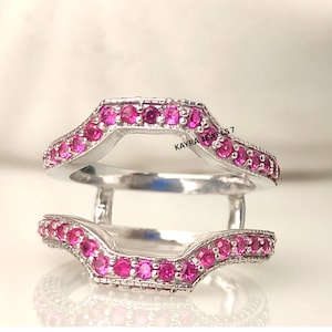 Puede incluir: Un protector de anillo plateado con un diseño curvo, engastado con numerosas piedras preciosas rosas redondas y pequeñas. El protector de anillo está diseñado para usarse con otro anillo. El texto "KAYRA JEWELS 7" es visible.