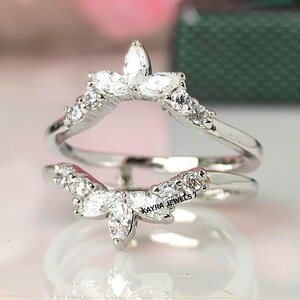 Puede incluir: Un protector de anillo plateado con un dise&ntilde;o curvo. El anillo presenta m&uacute;ltiples piedras claras y brillantes, incluyendo gemas de talla marquesa y redondas. El texto "KAYRA JEWELS 7" es visible en el anillo.