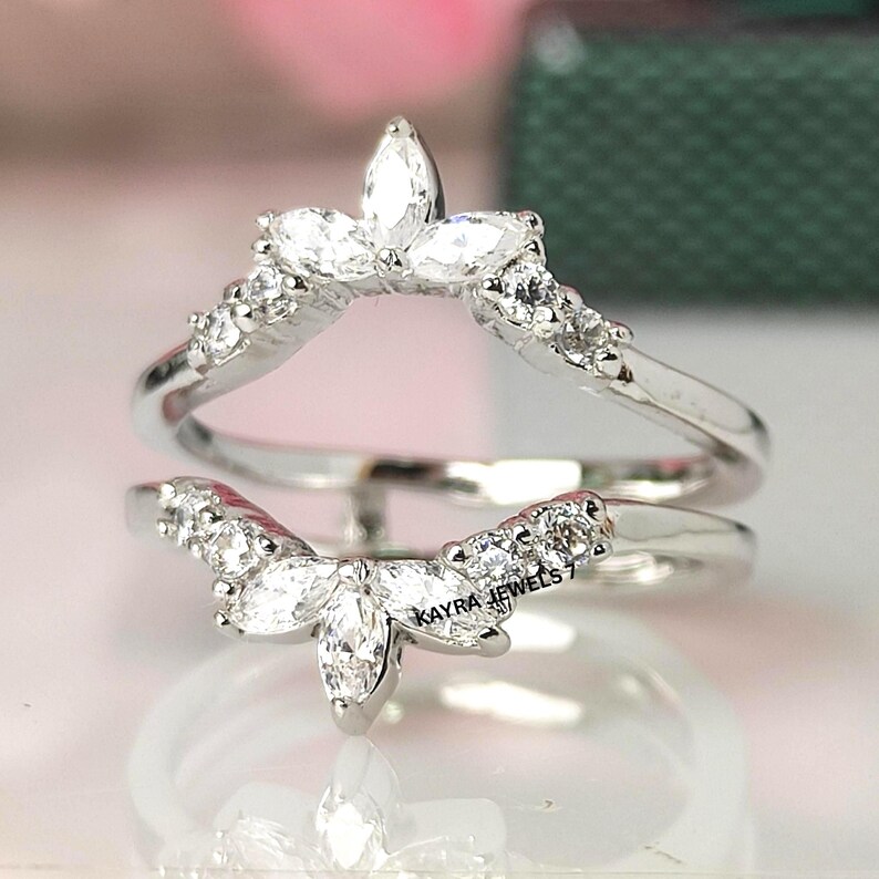 Puede incluir: Un protector de anillo plateado con un dise&ntilde;o curvo. El anillo presenta m&uacute;ltiples piedras claras y brillantes, incluyendo gemas de talla marquesa y redondas. El texto "KAYRA JEWELS 7" es visible en el anillo.