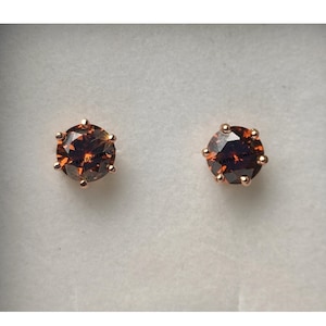 May include: A pair of stud earrings with round, faceted, orange-brown gemstones set in rose gold-coloured metal. The gemstones are held by six prongs. The earrings are displayed on a white background.