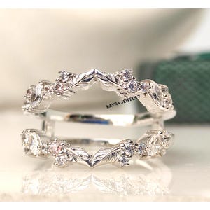 Puede incluir: Un anillo plateado con diseño de hojas, adornado con piedras preciosas claras y rosas. El anillo presenta un delicado patrón inspirado en la naturaleza con una forma de V central. El texto "KAYRA JEWELS7" es visible.
