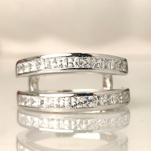 May include: A silver ring featuring two bands, each set with square-cut, clear gemstones. The ring has a contemporary, open design, with the bands connected by small supports. The text "KAYRA JEWELS 7" is visible on the ring.