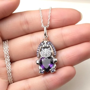 May include: A silver necklace featuring a pendant in the shape of a cartoon character. The pendant has a purple heart-shaped gemstone, framed by silver and smaller purple and white gemstones. The necklace is held in a hand.