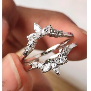 Puede incluir: Un anillo de plata con un diseño único, con piedras preciosas claras de talla marquesa y redonda. El anillo tiene una forma inspirada en flores, con las piedras preciosas dispuestas en una delicada disposición. El anillo es sostenido por una mano.