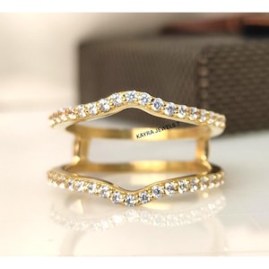 May include: A gold ring guard with a double band design. The bands are set with small, clear gemstones. The text "KAYRA JEWELS 7" is visible on the side of the ring.