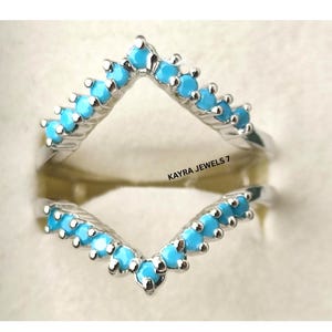 May include: A silver ring guard with a V-shaped design, adorned with small, round, light blue gemstones. The ring guard is designed to be worn with a solitaire ring. The text "KAYRA JEWELS 7" is visible.
