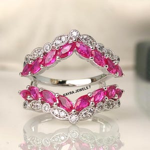 Puede incluir: Dos protectores de anillo plateados con piedras rosas talladas en marquesa y pequeños detalles blancos. Los anillos están apilados, creando un efecto en capas. El texto "KAYRA JEWELS 7" es visible.