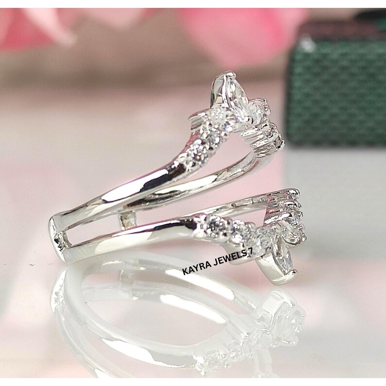 Puede incluir: Un protector de anillo plateado con piedras preciosas claras de talla marquesa y redonda. El protector de anillo tiene un dise&ntilde;o curvo y est&aacute; dise&ntilde;ado para usarse con un anillo solitario. El texto "KAYRA JEWELS_7" es visible.