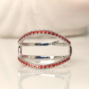 Pode incluir: Um anel de prata com duas faixas, cada uma com pequenas pedras vermelhas. O anel tem um design aberto único, com as faixas conectadas por barras verticais. O texto "925 KAYRA JEWELS 7" é visível no anel.