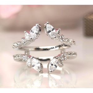 Puede incluir: Un par de realzadores de anillos plateados, cada uno con una fila de pequeñas piedras preciosas redondas y transparentes y una piedra preciosa transparente más grande con forma de marquesa. El texto "KAYRA JEWELS 7" es visible.