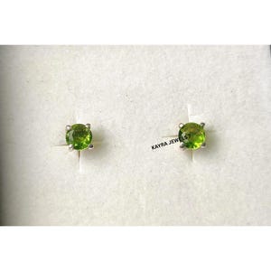 May include: Stud earrings with round, light green gemstones set in silver-coloured metal. The earrings are presented in a white jewellery box. The text "KAYRA JEWELS7" is visible.