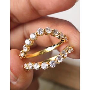 Puede incluir: Un anillo dorado con un diseño de doble banda, cada uno adornado con una fila de piedras preciosas redondas y transparentes. El anillo está sostenido por una mano, mostrando los intrincados detalles de la joya. El texto "KAYRA JEWELS" es visible.