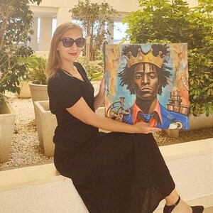 May include: An artwork featuring a portrait of a person wearing a gold crown, with a blue and orange background. The artwork is held by a person wearing a black dress and sunglasses.