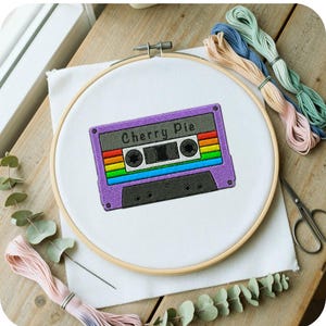 Könnte beinhalten: Ein runder hölzerner Stickrahmen mit weißem Stoff, der ein maschinengesticktes violettes Kassettendesign zeigt. Die Kassette hat einen Regenbogenstreifen, ein graues Etikett mit dem Text Cherry Pie und schwarze Details. Stickgarn liegt daneben.