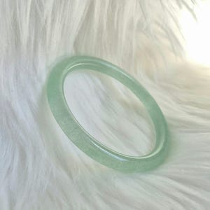 May include: A pale green jade bangle. The circular bracelet is a light green colour and sits on a white, fluffy surface. The bangle is a simple, elegant piece of jewellery, suitable for everyday wear.